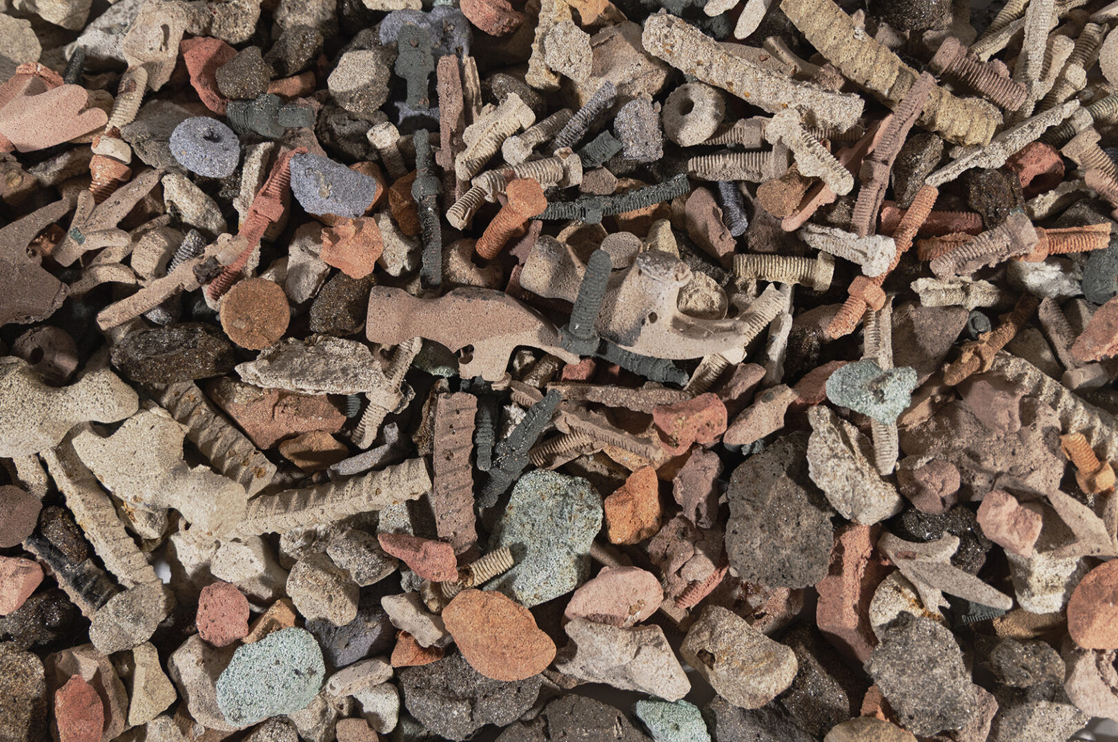 an overhead view of a colorful pile of rubble; hammer heads, rebar, screws and stones have been reproduced in a grainy material which looks like packed sand.