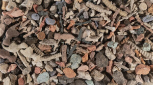 an overhead view of a colorful pile of rubble; hammer heads, rebar, screws and stones have been reproduced in a grainy material which looks like packed sand.