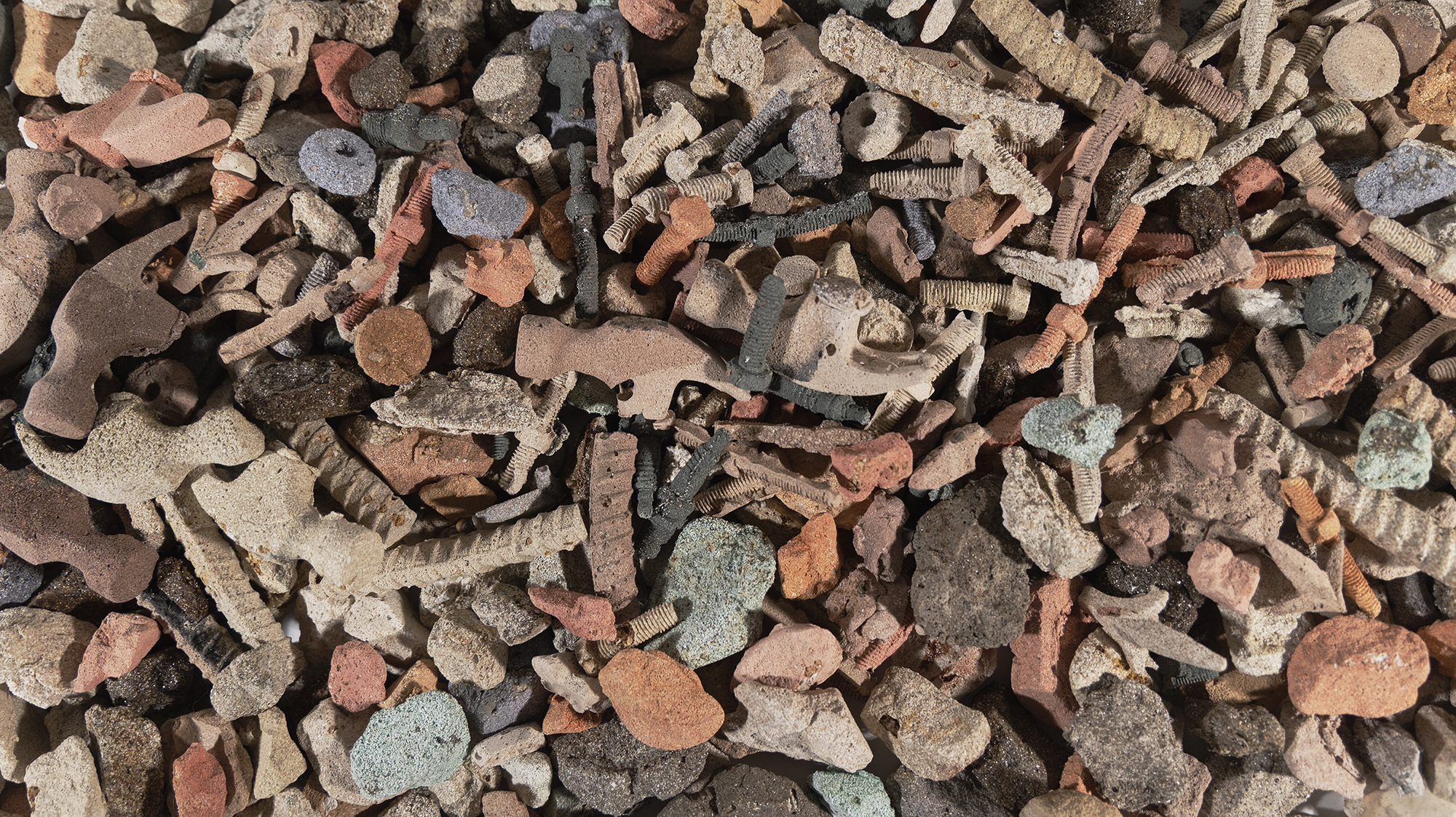an overhead view of a colorful pile of rubble; hammer heads, rebar, screws and stones have been reproduced in a grainy material which looks like packed sand.
